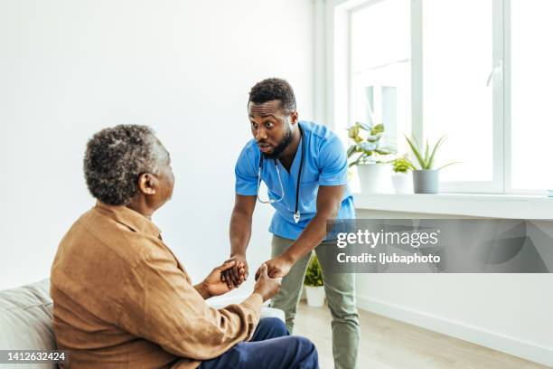 männlicher häuslicher betreuer, der den alten mann beim aufstehen vom sofa im pflegeheim unterstützt - nurse with old male patient home care stock-fotos und bilder