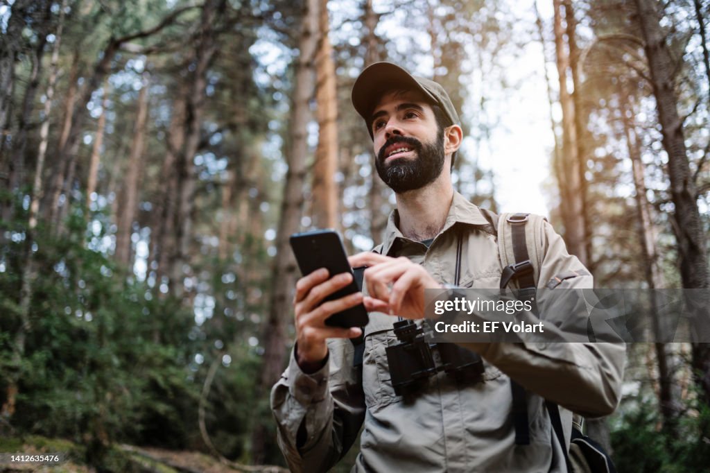 Young man hiker with beard and wearing a green cap and binoculars using cell phone app in the forest