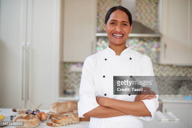a happy female baker standing arms crossed in a modern kitchen of a bakery with pastries in the background. a female chef wearing chef jacket or coat standing with arms folded and looking confident - chef coat stock pictures, royalty-free photos & images