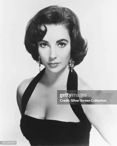 Elizabeth Taylor , British actress, wearing a black halterneck top and drop earrings, looking glamorous in a studio portrait, against a white...
