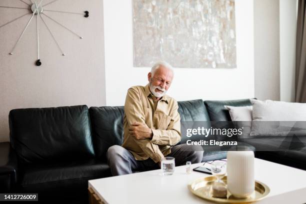 senior man rubbing his hands in discomfort, suffering from arthritis in his hand while sitting on sofa at home. elderly and health issues concept, pain in the hands can be symptoms of carpal tunnel - karpaltunnelsyndrom bildbanksfoton och bilder