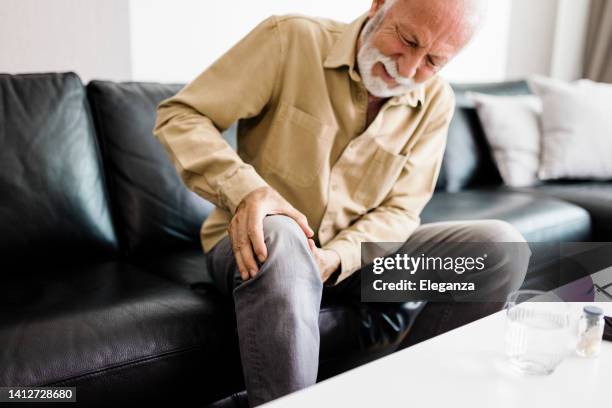 a distraught senior man feeling unwell, suffering from pain in leg while sitting on sofa in the living room at home, photo of elderly man sitting on a sofa in the living room at home and touching his knee by the pain during the day. senior man massaging - knee stock pictures, royalty-free photos & images