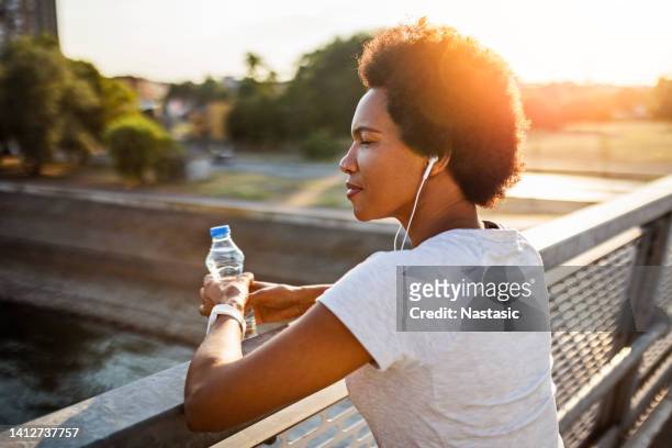 young woman taking a break from jogging across the bridge at sunset - woman headphones sport smile iphone stock pictures, royalty-free photos & images