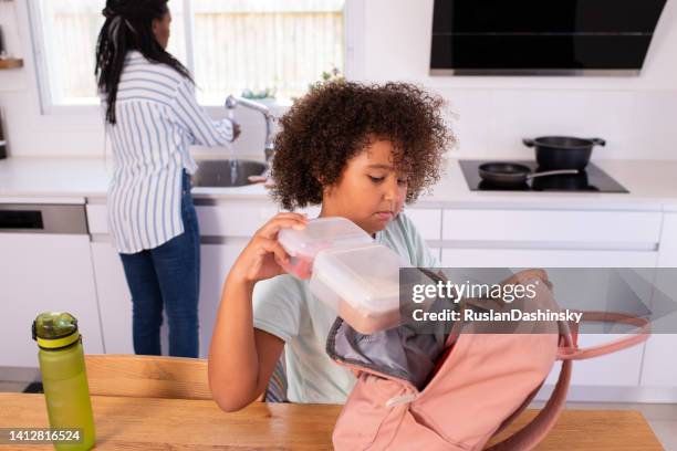 an elementary-age curly hair kid putting a full lunch box into a pink backpack before school. - open rucksack stock pictures, royalty-free photos & images