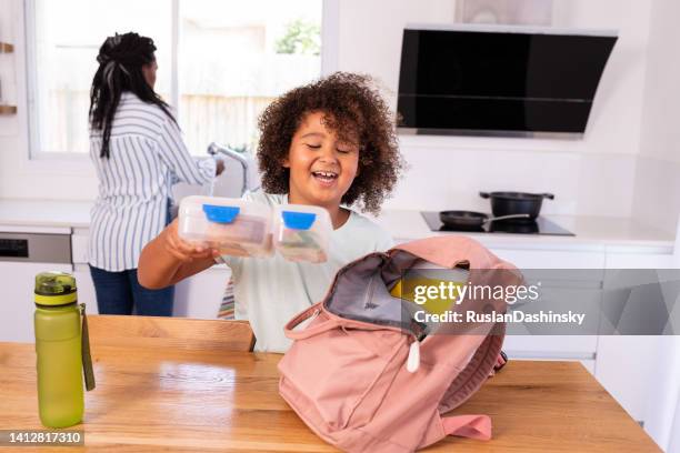 happy elementary-age child putting a lunch box into a pink backpack before going to school. - packing backpack stock pictures, royalty-free photos & images