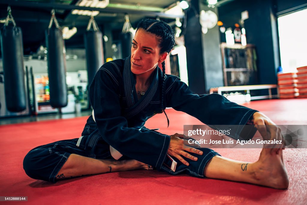 Woman in black karategi stretching her legs in a training gym, Sporty woman in black kimono practicing leg stretching before starting karate fight in the gym.
