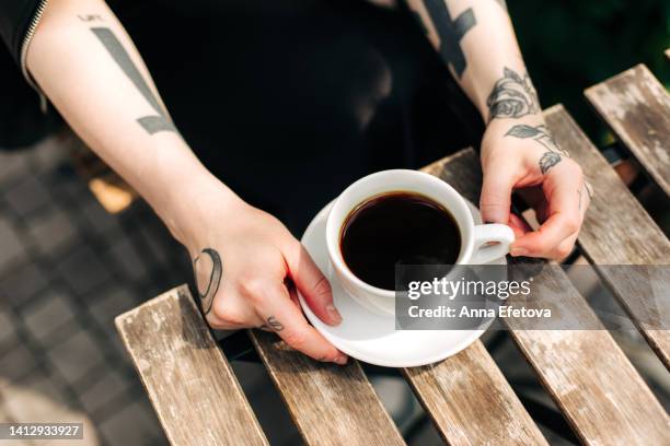 female hands with many tattoos are holding a white cup of a black coffee on a wooden table. concept of enjoying morning hours before a working day - café preto imagens e fotografias de stock