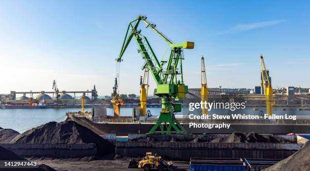 large green dockside crane loads coal into cargo hold of a freight ship in gdansk harbor with piles of coal and two bulldozers in the foreground - poland sea stock pictures, royalty-free photos & images