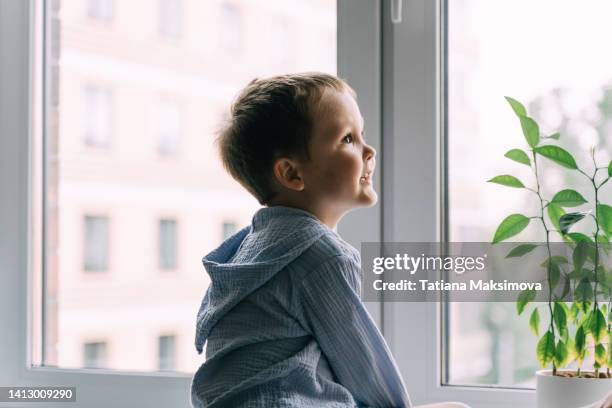 a little boy is sitting on the windowsill against the background of the window, profile view. - windowsill copy space stock pictures, royalty-free photos & images