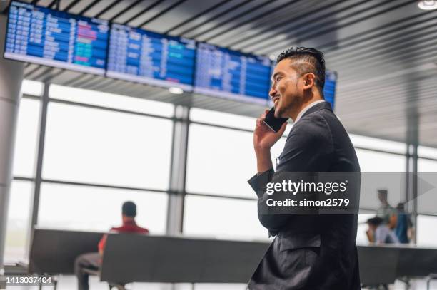 an asian businessman standing in front of departure board using mobile phone, travel, information, connected - departure board front on stockfoto's en -beelden