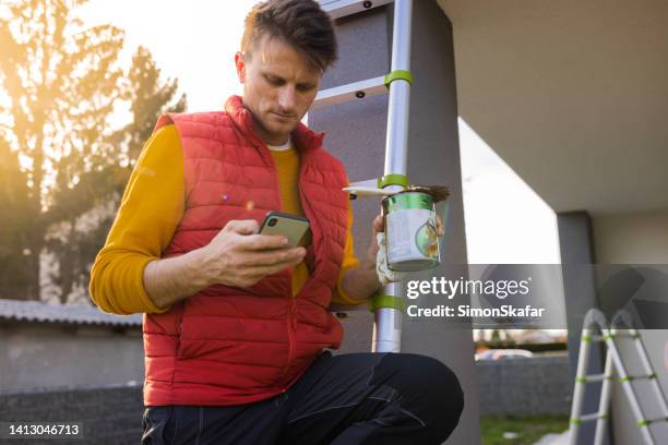 man holding paint equipment while using smart phone on ladder outside apartment - backyard renovation stock pictures, royalty-free photos & images