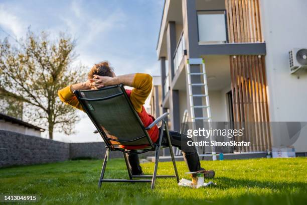 tired man resting on deck chair by paint equipment in yard outside apartment - backyard deck stockfoto's en -beelden