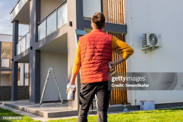 rear view of man with paintbrush and paintcan looking at apartment from yard - backyard renovation stock pictures, royalty-free photos & images
