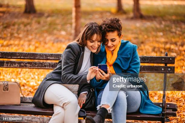 two young women using phone on a bench in the park in autumn - autumn friends coats stock pictures, royalty-free photos & images