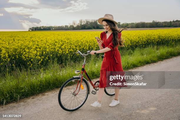 woman using cellphone while walking with bicycle on road against canola field - hungary countryside stock pictures, royalty-free photos & images