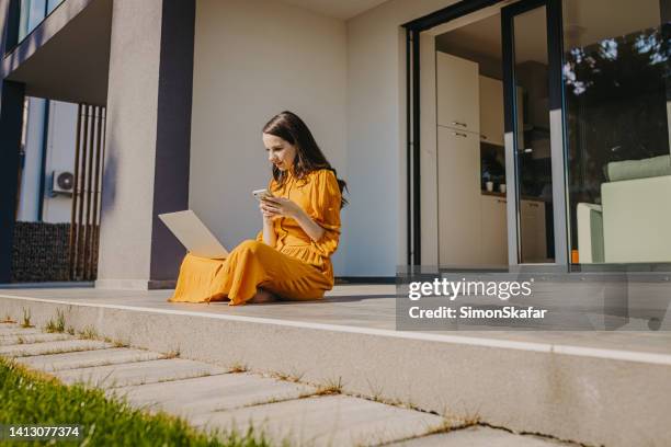 mujer con computadora portátil usando el teléfono celular mientras está sentada en el porche durante el día soleado - columnista fotografías e imágenes de stock