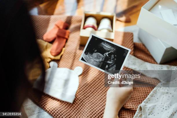 over the shoulder view of pregnant woman looking at an ultrasound scan photo, with baby clothings and accessories laying on the floor against sunlight. mother-to-be. preparation for a new family member. expecting a new life concept - fetal position fotografías e imágenes de stock