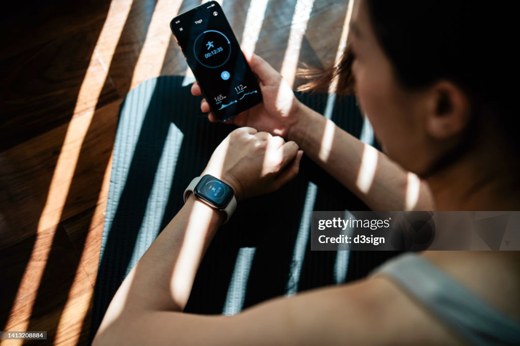 Young active Asian woman exercising at home, using smartphone health and fitness apps to monitor and track training performance and activity. Recording health status with mobile phone. Health and fitness training with technology. Healthy living lifestyle