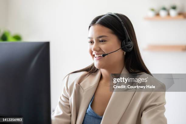 young woman working in call center takes calls on headset while looking at computer screen - filipino women stock pictures, royalty-free photos & images