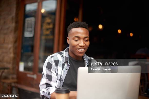 trendige und stilvolle junge schwarze studentin, die auf einem laptop im freien in einem café in der stadt tippt. glücklicher afrikanischer mann, der lächelt, während er vor einem restaurant studiert und ein projekt schreibt - african creative with laptop working outside stock-fotos und bilder