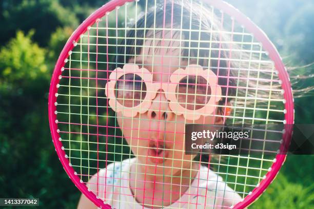 funny little asian girl wearing flower-shaped sunglasses making a silly face behind badminton racket, having fun playing in the park. enjoying nature and summer days outdoors. outdoor fun. childhood lifestyle - playing badminton stock-fotos und bilder