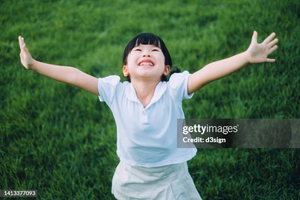 happy little asian school girl having fun playing on the meadow in park. smiling joyfully with her arms outstretched. enjoying nature and free time on summer day outdoors. childhood lifestyle. carefree, freedom and playful concept - student achievement stock pictures, royalty-free photos & images