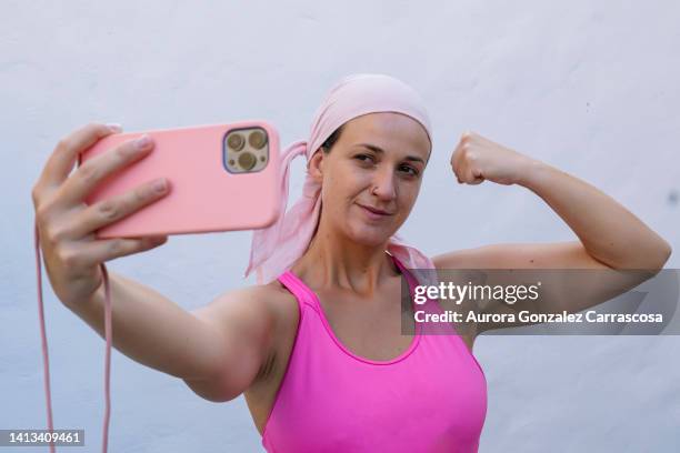 woman using smartphone to take a selfie, wearing a pink head covering. in concept of fight against cancer. - fighting cancer stock pictures, royalty-free photos & images