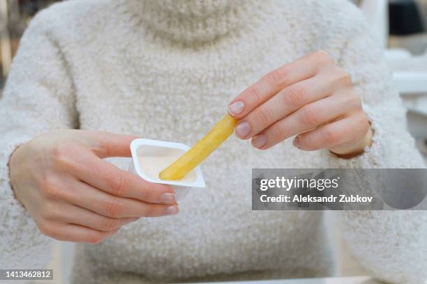 a woman holds french fries in her hand while eating them in a cafe or restaurant. a girl in a white knitted sweater dips a slice of potato in garlic sauce. the concept of fast food, snacks, junk food. food and beverage establishment. - plump girls stockfoto's en -beelden