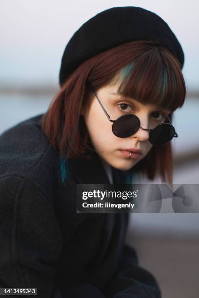 fille rousse vêtue de noir sur fond de rivière à l’extérieur. style gothique. modèle de mode asiatique en manteau sombre avec parapluie sur nature. lunettes de soleil noires. tenue élégante. style moderne de la jeunesse. sous-culture - goth photos et images de collection