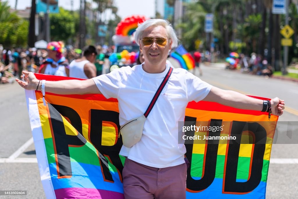 Lächelnder schwuler Mann im mittleren Erwachsenen, der ein Regenbogenbanner hält, das das Wort stolz in einer Pride-Parade zeigt