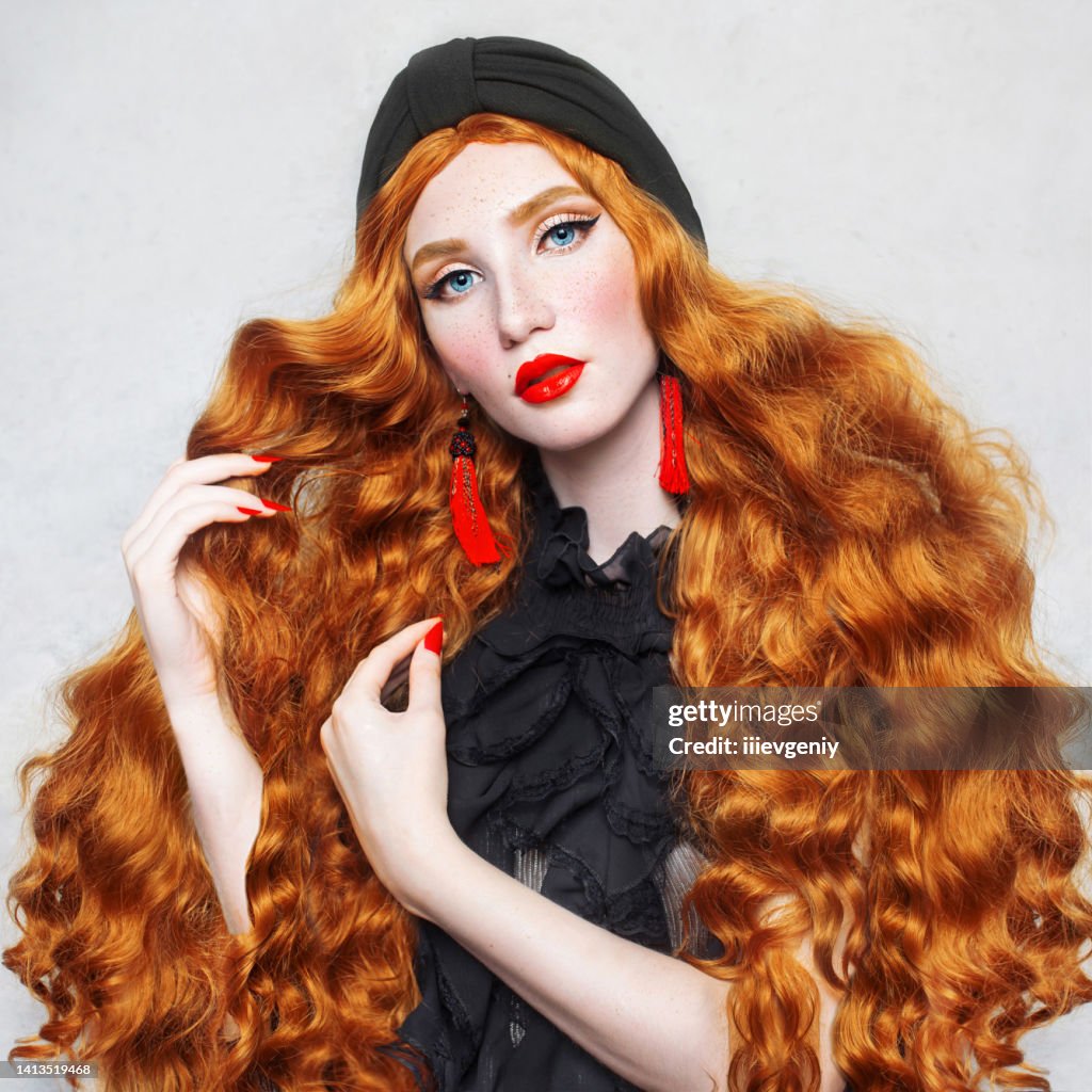 Redhead fabulous woman. Long curly hair. Beauty makeup. Pale skin. Girl in turban and black dress on white background. Beautiful model with red lips. Retro outfit. Hairstyle. Jewelry and bijouterie