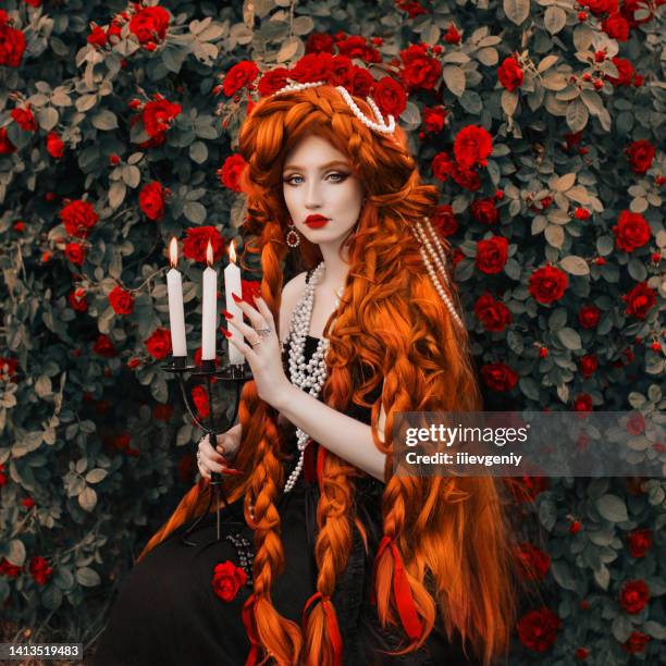 redhead vampire woman. long curly hair. pale skin. girl in black dress on rose flower background. beautiful model with red lips. renaissance outfit for halloween. hairstyle. jewelry and bijouterie - long necklace stockfoto's en -beelden