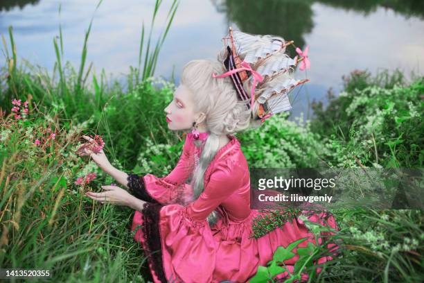 renaissance princess with blonde hair on lake background. beauty makeup. fairytale rococo queen with ship in hairstyle on nature. model in pink dress. woman with historical hair style sit in grass. - female model hair bun stock pictures, royalty-free photos & images