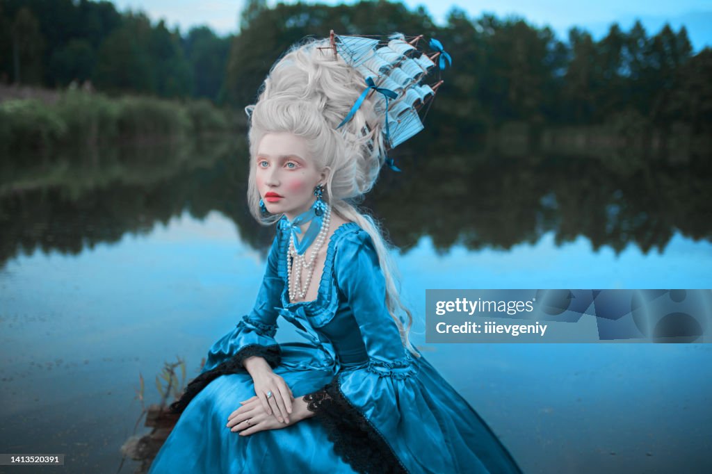Renaissance princess with blonde hair on lake background. Beauty makeup. Fairytale rococo queen with ship in hairstyle on nature. Model in blue dress. Woman with historical hair style on bridge