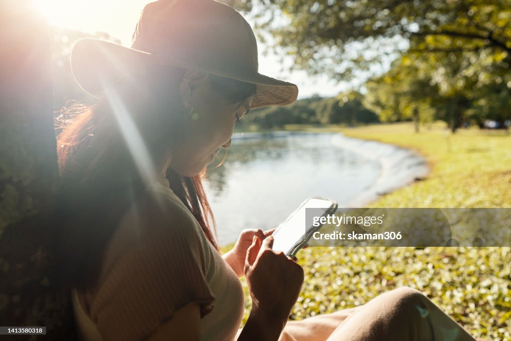 Positive emotion Asian teenager Gen z sitting under the tree leisure public park and using phone to take a photo for a post on the social media platform.