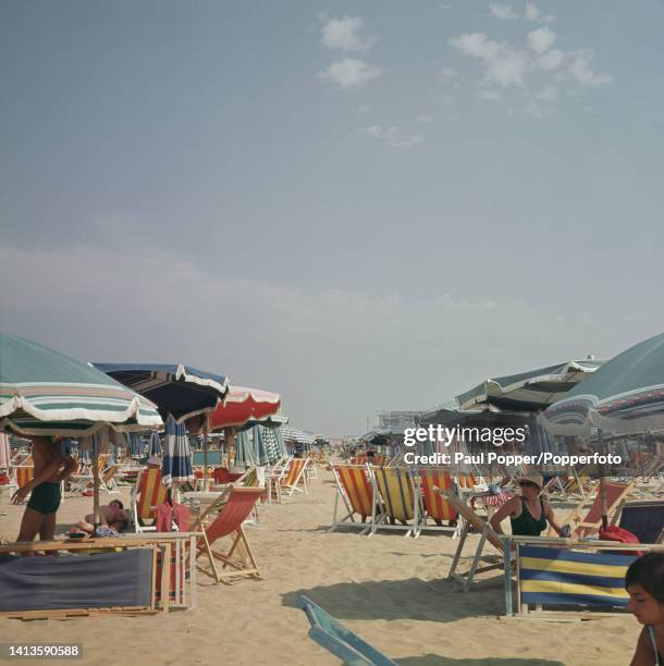 Holidaymakers and visitors seated on deck chairs under umbrellas and parasols on the sandy beach at Rimini on the Adriatic coast of the...