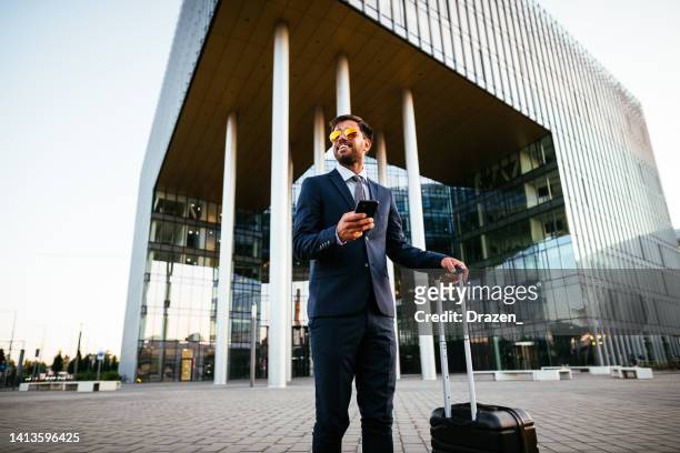 businessman in full suit near modern office building or airport, with suitcase and sunglasses - hungary hotel stock pictures, royalty-free photos & images