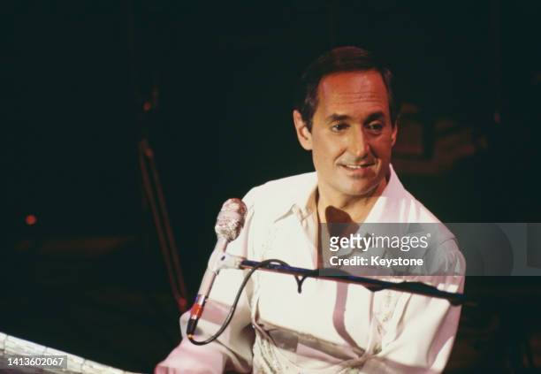 American singer, songwriter and pianist Neil Sedaka sitting at a piano as he performs live in concert at a venue, 1986.