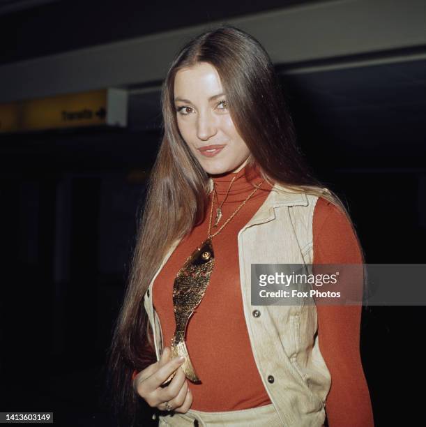 British actress Jane Seymour, wearing a corduroy waistcoat over a red turtleneck sweater, holding the tail of the large gold fish pendant hanging...