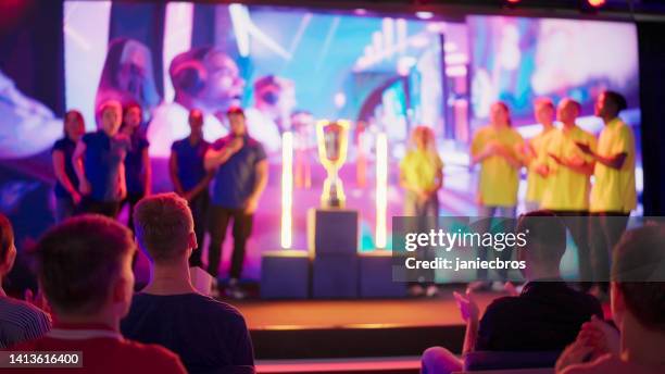 diverse yellow and blue pro gamers team with african ethnicity players standing on stage. trophy between them. esport championship competition - awards broadcast stock pictures, royalty-free photos & images