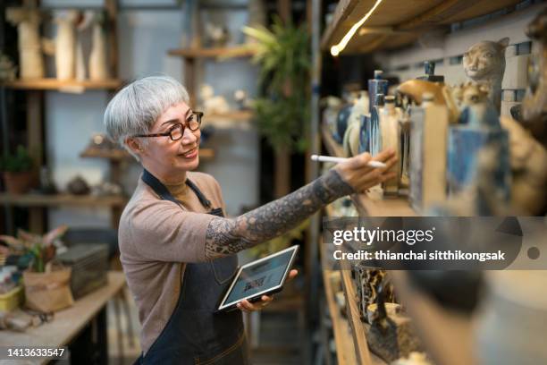 senior artist woman using a digital tablet examining pottery in her studio. - kleinunternehmen stock-fotos und bilder