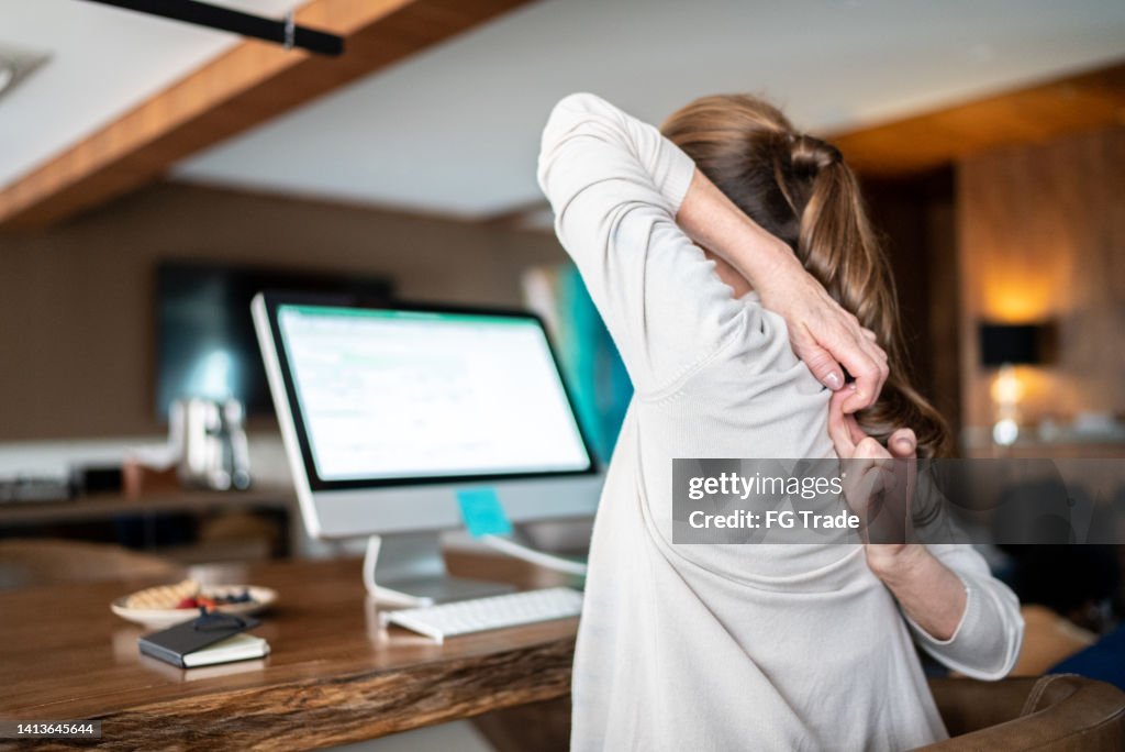 Reife Frau streckt sich auf dem Stuhl, während sie den Computer zu Hause benutzt