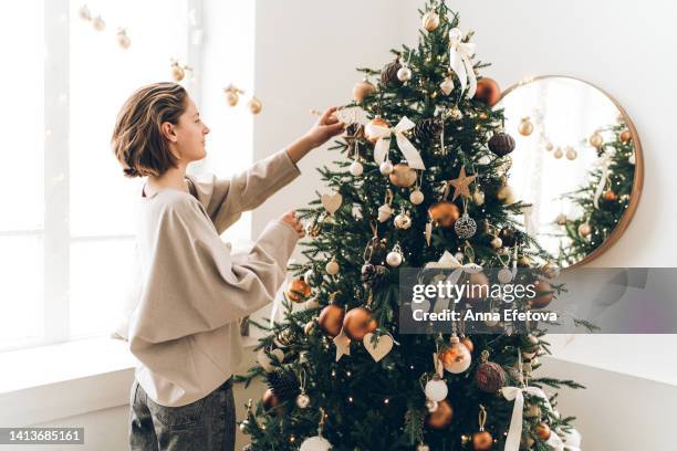 young woman is adjusting her hair and decorating christmas tree with many different decorations and festive garland. new year celebration concept - decorare l'albero di natale foto e immagini stock