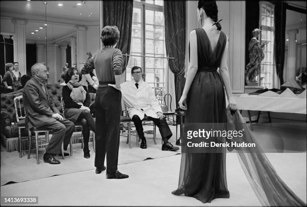 View of French fashion designer Yves Saint Laurent as he and others examine an outfit on an unidentified model during the preparations for a haute...