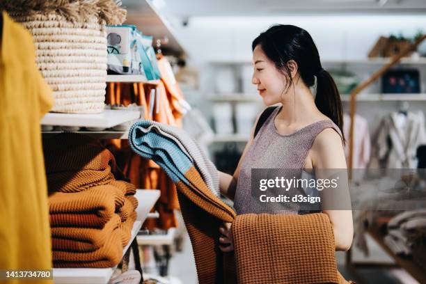 smiling young asian woman shopping for home decor and necessities in a homeware store, choosing for a blanket on a shelf - comprando - fotografias e filmes do acervo