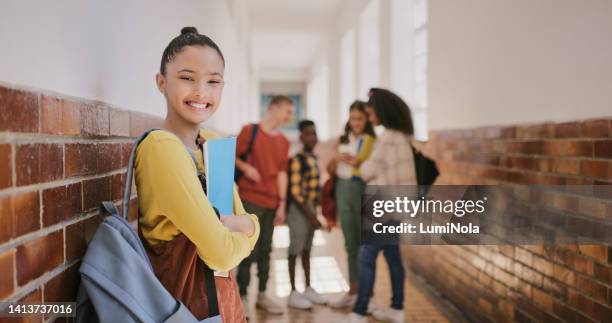 cute young girl excited to be at school and learning with her friends holding her education notebook bag. portrait of smiling student standing in the hallway with her class mates in the background - scholarship award stock pictures, royalty-free photos & images