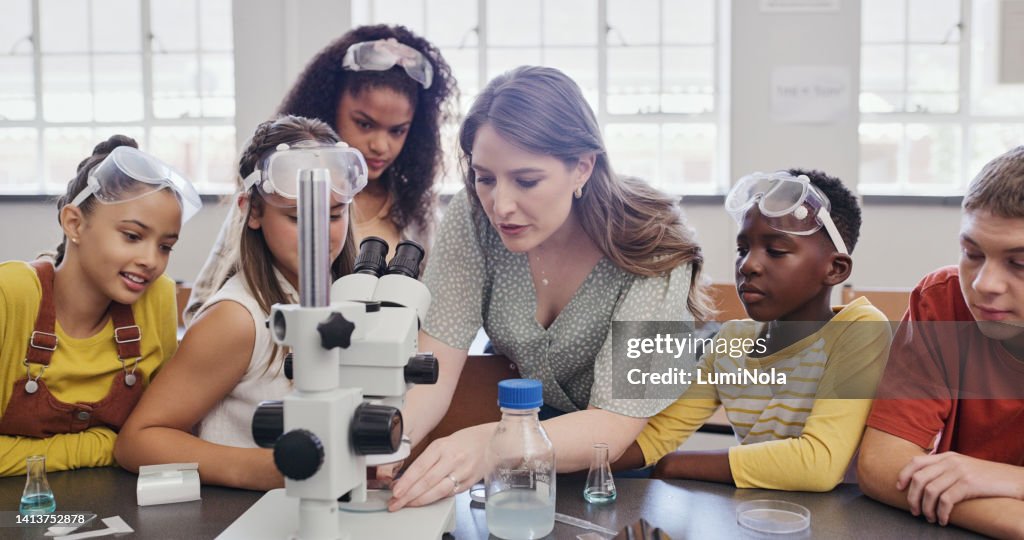 Female science teacher explaining to a classroom of young students. Boys and girls doing an experiment with adult supervision and examining substances under a microscope at school