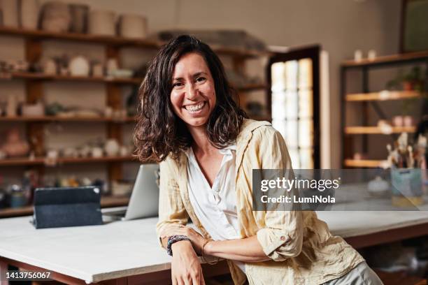 portrait of a happy, smiling and laughing female business owner leaning on the desk of her art studio. a creative and relaxed female looking at the camera with a blurred background of a ceramic store - portrait blurred background stockfoto's en -beelden