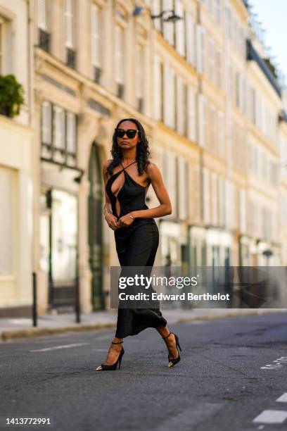 Emilie Joseph @in_fashionwetrust wears black sunglasses, a black satin halter-neck / V-neck / cut-out chest long dress, black shiny leather pointed...