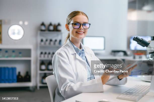 professionelle, lächelnde und glückliche wissenschaftliche ärztin in einem modernen medizinlabor mit einem mikroskop. ein porträt einer frau, die an ihrem schreibtisch sitzt und für die kamera lächelt, während sie auf ihr tablet drückt. - female pharmacist with a digital tablet stock-fotos und bilder